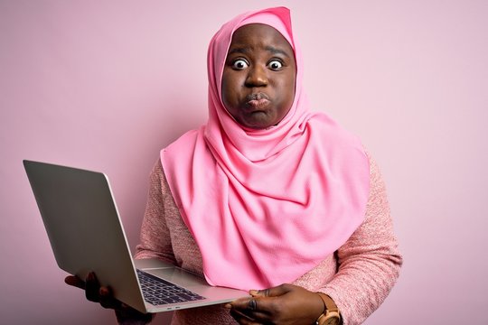 African american plus size woman wearing muslim hijab using laptop over pink background puffing cheeks with funny face. Mouth inflated with air, crazy expression.