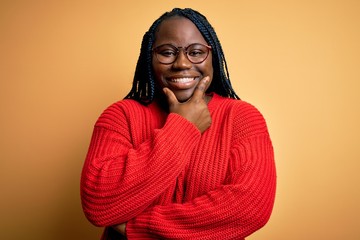 African american plus size woman with braids wearing casual sweater over yellow background looking confident at the camera smiling with crossed arms and hand raised on chin. Thinking positive.