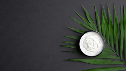 Jar on natural cosmetic cream moisturizer and palm leaf on black background. Flat lay, top view. SPA organic beauty product, skincare concept.