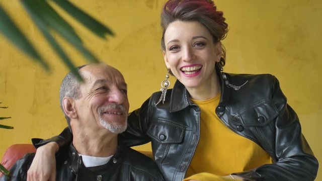 Emotional family portrait of adult daughter and senior father in loft room wearing black leather jackets in punk style and looking to each other face, girl is showing rock and roll gesture.