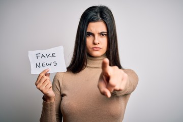 Young beautiful brunette woman holding paper with fake news message over white background pointing...