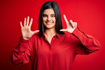 Young beautiful brunette woman wearing casual shirt standing over red background showing and pointing up with fingers number seven while smiling confident and happy.