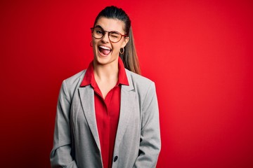 Young beautiful brunette businesswoman wearing jacket and glasses over red background winking...