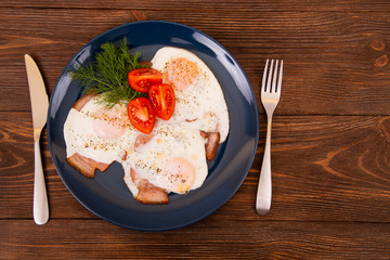 Dark plate with fried eggs, pork ham, dill and tomatoes on wooden textured background, fork and knife, top view