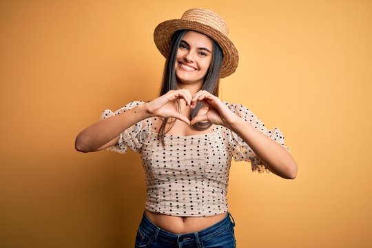 Young beautiful brunette woman wearing t-shirt and summer hat over yellow background smiling in love showing heart symbol and shape with hands. Romantic concept.