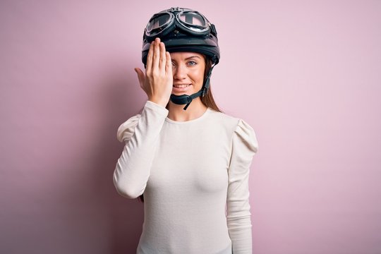 Young beautiful motorcyclist woman with blue eyes wearing moto helmet over pink background covering one eye with hand, confident smile on face and surprise emotion.