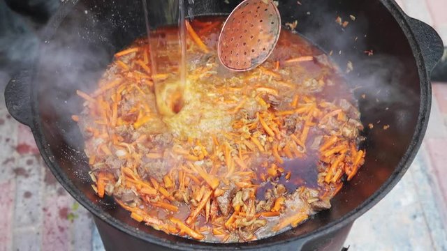 Preparation of national uzbek dish pilaf, pilaw, plov, carrot with meat in big pan. Add water, Cooking process, open fire. Cooking in a cauldron on fire. Stir slowly with a skimmer. Add spices