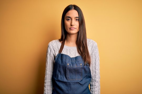 Young beautiful baker woman wearing apron uniform cooking over yellow background with serious expression on face. Simple and natural looking at the camera.