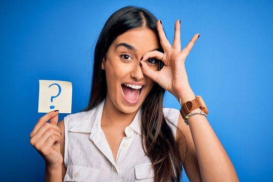 Young beautiful brunette woman holding paper with question mark symbol message with happy face smiling doing ok sign with hand on eye looking through fingers