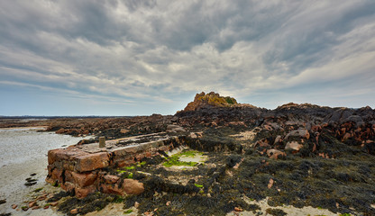 Imge of the beach at low tide with rocks, seaweed, small stone pier and a cloudy sky at Le Hocq, St Clements, Jersey Channel Islands.