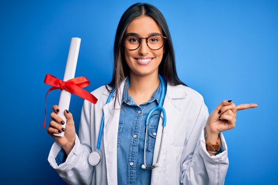 Young beautiful brunette doctor woman wearing glasses and coat holding diploma degree very happy pointing with hand and finger to the side