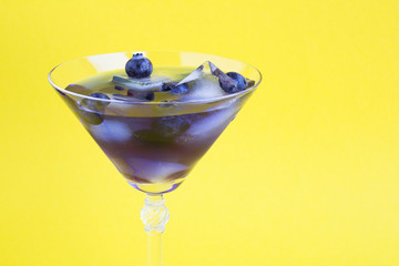 Blue cocktail drink with ice and blueberries in a martini glass on the yellow  background. Closeup. Copy space.