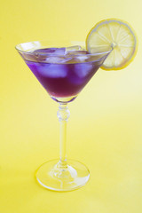 Iced violet drink or cocktail with lemon in a martini  glass on the yellow background. Location vertical. Closeup.