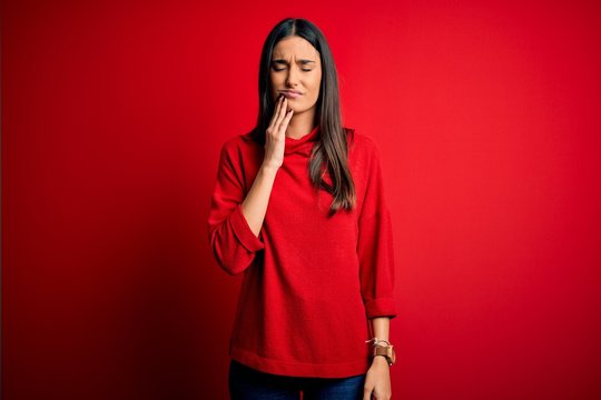 Young beautiful brunette woman wearing casual sweater over isolated red background touching mouth with hand with painful expression because of toothache or dental illness on teeth. Dentist
