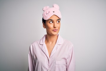 Young beautiful brunette woman wearing pajama and sleep mask over white background smiling looking to the side and staring away thinking.