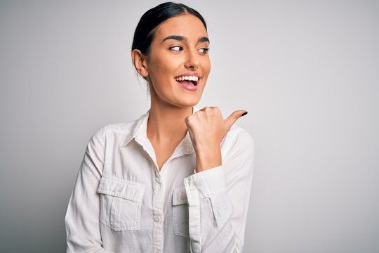 Young beautiful brunette woman wearing casual shirt over isolated white background smiling with happy face looking and pointing to the side with thumb up.