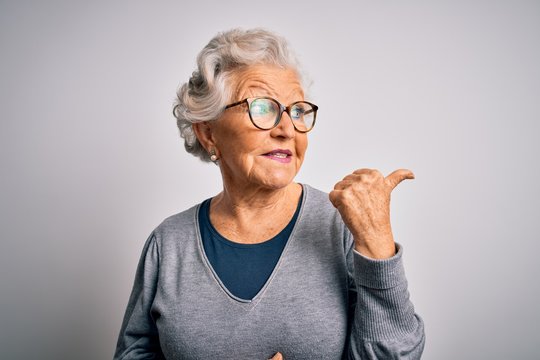 Senior beautiful grey-haired woman wearing casual sweater and glasses over white background smiling with happy face looking and pointing to the side with thumb up.