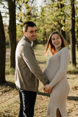 Obraz premium happy young pregnant couple walks in the park,husband holds the hand of his pregnant wife,future dad and mom,walks in the park after quarantine