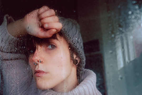 Unhappy pensive young female in warm sweater and hat looking away through steamy window while spending day at home in self isolation