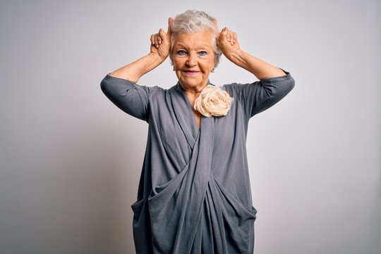 Senior beautiful grey-haired woman wearing casual dress standing over white background doing funny gesture with finger over head as bull horns