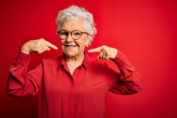Senior beautiful grey-haired woman wearing casual shirt and glasses over red background smiling...