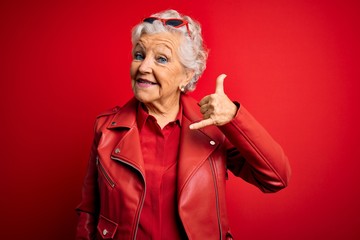 Senior beautiful grey-haired woman wearing casual red jacket and sunglasses smiling doing phone...