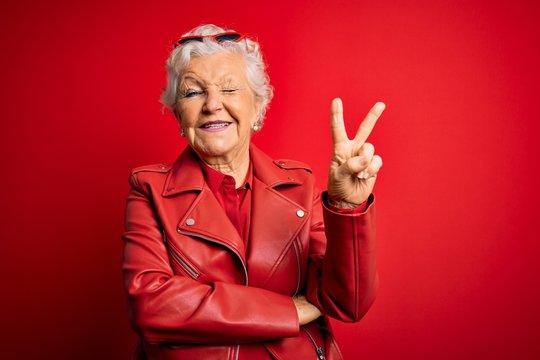 Senior beautiful grey-haired woman wearing casual red jacket and sunglasses smiling with happy face winking at the camera doing victory sign. Number two.