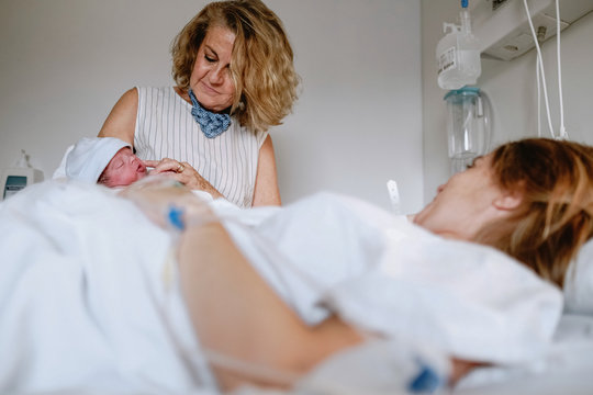Happy mature female greeting newborn grandchild in hospital ward while young mother lying in bed and receiving medical treatment