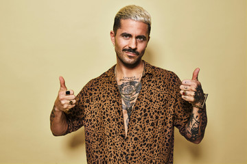 Positive young bearded guy in stylish leopard shirt revealing tattooed torso showing thumbs up gesture while standing against beige background