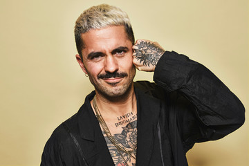 Portrait of unfriendly arrogant stylish man with muscular tattooed torso wearing black coat looking at camera against beige background making hand punch gesture to his temple