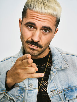Young ethnic bearded man with trendy hairdo dressed in denim jacket pointing at camera with challenge while standing against gray background