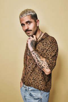 Fashionable male model with tattoos wearing trendy leopard shirt and jeans standing against beige background and looking at camera
