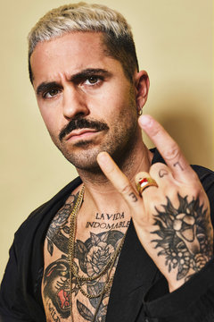 Young provocative male with mustache in black coat over naked tattooed torso showing rock sign gesture while standing against beige background looking at camera