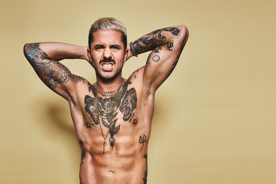 Handsome sexy attractive muscular male with various tattoos on naked torso and arms looking at camera making faces with mouth while standing against beige background