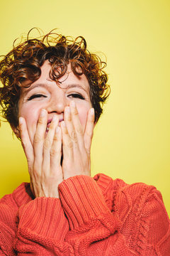 Amazed laughing female with curly hair wearing orange knitted sweater covering mouth with hands and looking away while standing against yellow background