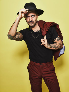 Handsome well dressed male with mustache in trendy hat and vinous suit holding suit coat over the shoulder looking at camera while standing against yellow background