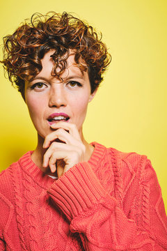 Amazed female with curly hair wearing orange knitted sweater with hand on mouth looking at camera while standing against yellow background