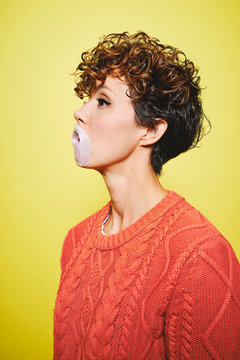 Side view of young pretty curly haired female in orange sweater trying to blow a bubble gum while standing looking away against yellow background