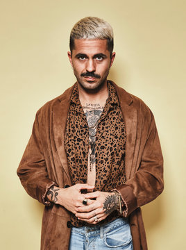 Fashionable Male Model With Tattoos Wearing Trendy Coat Over Leopard Shirt And Jeans Standing Against Beige Background And Looking At Camera