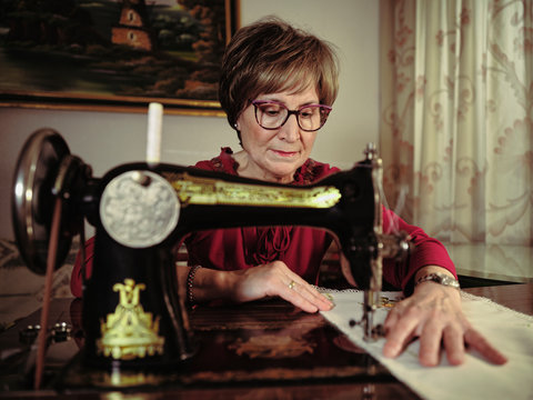 Senior lady in glasses using retro sewing machine to create linen napkin in cozy room at home