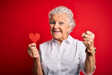 Senior beautiful woman holding paper heart standing over isolated red background screaming proud...
