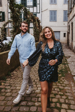 Cheerful young couple in stylish casual clothes holding hands and smiling while walking on old narrow street in city