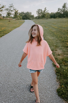 Cheerful preteen girl in casual hoodie shirt and shorts looking away while walking on pathway in park in summer day