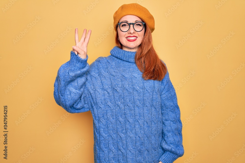 Sticker Young beautiful redhead woman wearing french beret and glasses over yellow background showing and pointing up with fingers number three while smiling confident and happy.