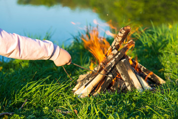 Children's hand sticks a wooden branch in a burning bonfire by the river in the summer