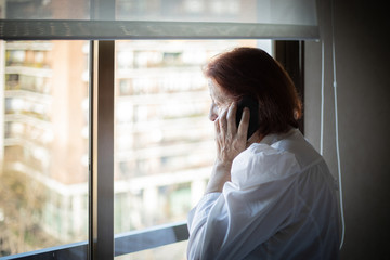 Side view of senior female in white blouse having phone call while standing near window in hospital ward visiting relative