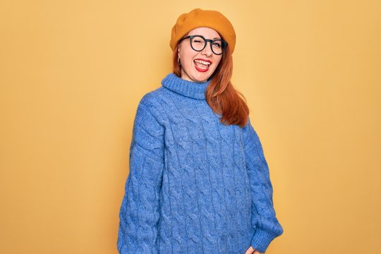 Young beautiful redhead woman wearing french beret and glasses over yellow background winking looking at the camera with sexy expression, cheerful and happy face.