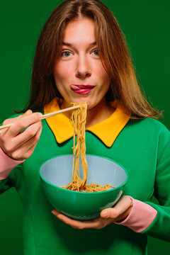 Naklejki Positive young female in green shirt looking at camera grimacing sticking tongue out while eating yummy instant noodles with chopsticks on green background
