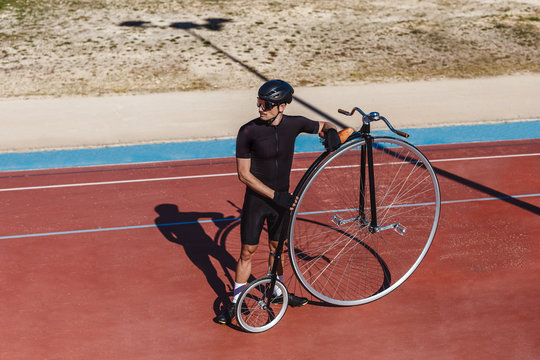 From above serious man in black activewear and helmet looking away while standing with retro penny farthing bicycle on racetrack at sports ground