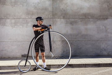 Calm determined man in black activewear and helmet looking away while standing on asphalt road and leaning on high wheel bicycle against concrete wall in sunny day